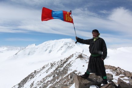 Ein Mann in traditioneller mongolischer Kleidung steht auf einem Felsen und hält die mongolische Flagge hoch in den Bergen.