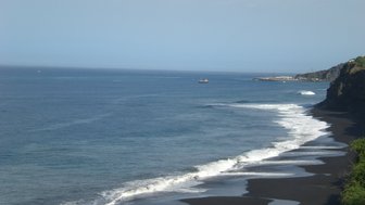Eine ruhige Küstenlandschaft mit schwarzem Sand, Wellen, die sanft an den Strand rollen, und einem malerischen Boot auf dem Wasser.