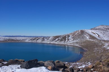 Ein klarer, blauer See umgeben von schneebedeckten Hügeln und einem strahlend blauen Himmel im Tongariro Nationalpark.