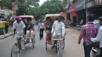 Zwei Rikschafahrer transportieren Touristen auf einer belebten Straße in Delhi, umgeben von Geschäften und Bäumen.