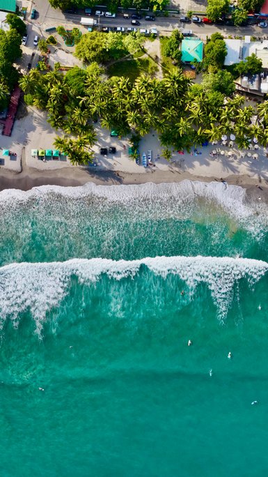 Eine luftige Perspektive zeigt den langen, ruhigen Strand mit palmengesäumter Küste und den sanft brechenden Wellen.