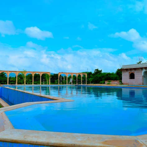 Ein weitläufiger Swimmingpool mit glitzerndem Wasser, umgeben von eleganten Bögen und üppigem Grün unter einem strahlend blauen Himmel.