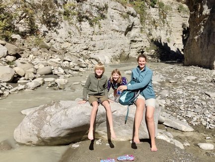 Drei Kinder und eine Frau sitzen auf einem großen Stein am Ufer eines schmalen Flusses, umgeben von steilen Felsen.