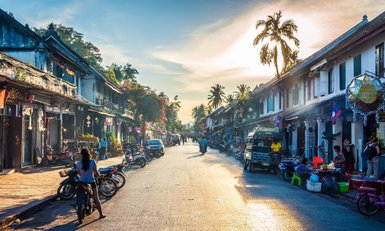 Eine belebte Straße in Luang Prabang, gesäumt von Geschäften und Motorrädern, während die Sonne am Horizont untergeht.
