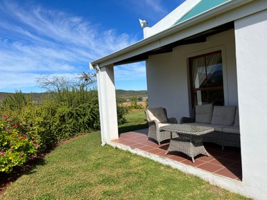 Eine gemütliche Terrasse mit einem Rattansofa und einem Tisch, umgeben von üppigem Grün und blühenden Pflanzen.
