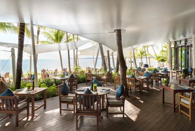 Ein elegantes Restaurant am Strand mit Holztischen und Stühlen, umgeben von Palmen und Blick auf das glitzernde Wasser.