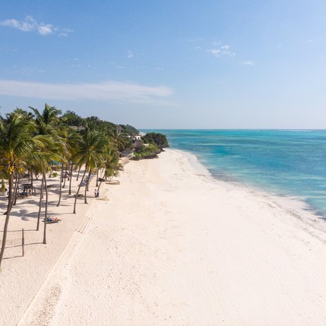 Ein weitläufiger, weißer Sandstrand mit Palmen, der sanft ins türkisfarbene Wasser übergeht, unter einem klaren blauen Himmel.