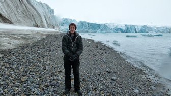 Ein Mann steht am Ufer eines kalten Gewässers, umgeben von Gletschern und schroffen Felsen in einer frostigen Landschaft.