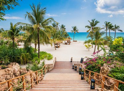 Eine malerische Treppe führt hinunter zu einem weißen Sandstrand, umgeben von Palmen und bunten Blumen.
