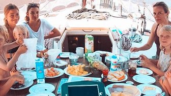 Eine fröhliche Gruppe von Kindern und Erwachsenen genießt ein gemeinsames Frühstück auf einem Segelboot, umgeben von Wasser.