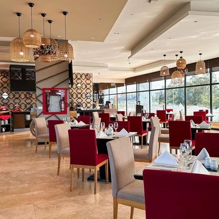 Ein elegantes Restaurant mit roten und beigen Stühlen, großen Fenstern und stilvollen Lampen, die eine einladende Atmosphäre schaffen.