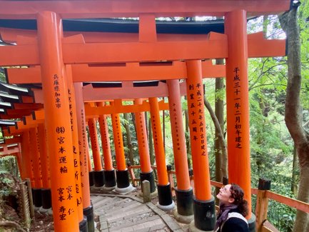Eine Person sitzt auf einer Steinbank und blickt lächelnd zu den leuchtend orangefarbenen Torii-Toren auf einem schmalen Weg.