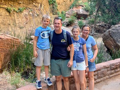 Eine Familie steht lächelnd vor einer beeindruckenden Felswand im Zion Nationalpark, umgeben von üppiger Vegetation.