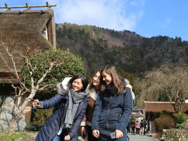 Drei junge Frauen lächeln und posieren für ein Selfie vor einem traditionellen japanischen Haus in einer malerischen Landschaft.