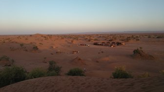 Eine weite Wüstenlandschaft mit sanften Sanddünen und einem kleinen Camp aus Zelten im Hintergrund, umgeben von spärlicher Vegetation.