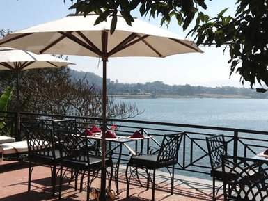 Ein malerischer Blick auf einen ruhigen See, umgeben von Bäumen und einem einladenden Restaurantbereich mit Sonnenschirmen.