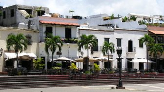 Blick auf die kolonialen Gebäude in Santo Domingo, umgeben von Palmen und einem einladenden Platz mit Tischen.