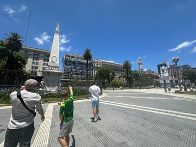 Ein Mann und ein Junge zeigen auf ein Denkmal in Buenos Aires, umgeben von modernen Gebäuden und Palmen unter blauem Himmel.