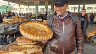Ein Mann in einer Markthalle lächelt, während er ein großes, frisch gebackenes Brot mit Sesam in der Hand hält.