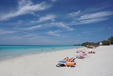 Ein sonniger Strand mit weißen Sandflächen, Liegen und Menschen, die im klaren Wasser schwimmen und sich entspannen.