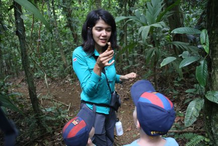 Eine Frau in einem grünen Hemd zeigt begeistert ein kleines Tier in der Hand, während Kinder aufmerksam zuhören.