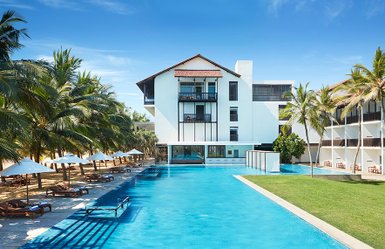 Ein modernes Hotelgebäude mit einem großen Pool, umgeben von Palmen und Liegen, strahlt tropische Entspannung aus.