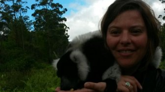 Eine Frau lächelt, während sie einen Lemur in ihrer Hand hält, umgeben von üppiger grüner Natur und blauem Himmel.