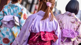 Drei Frauen in traditionellen Kimonos gehen durch eine belebte Straße, ihre bunten Rückansichten sind gut sichtbar.