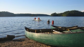 Zwei Personen stehen im Wasser, während ein Kanu mit Paddlern im Hintergrund über den ruhigen See gleitet.