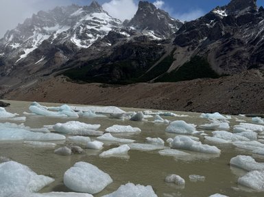 Im Vordergrund schwimmen Eisblöcke in einem trüben Gewässer, während majestätische Berge im Hintergrund aufragen.