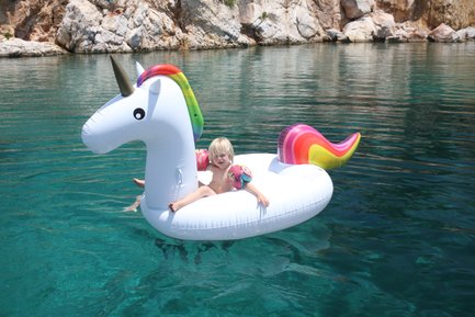 Ein kleines Kind sitzt auf einem großen, aufblasbaren Einhorn im klaren Wasser, umgeben von Felsen und Sonne.