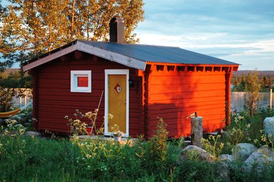 Ein rotes Holzhaus mit einem gelben Eingang und einem kleinen Fenster, umgeben von grünen Wiesen und Bäumen im Hintergrund.