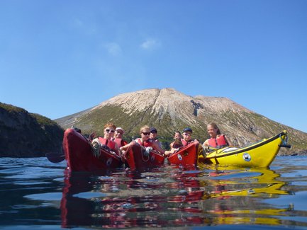 Sechs Kajakfahrer paddeln in ruhigem Wasser, während ein majestätischer Vulkan im Hintergrund thront.