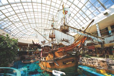 Ein großes, detailreiches Piratenschiff steht in einem Einkaufszentrum unter einem gläsernen Dach, umgeben von Wasser und Pflanzen.