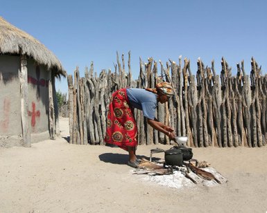 Eine Frau in traditioneller Kleidung kocht am Lagerfeuer vor einer hölzernen Umzäunung in einer ländlichen Umgebung.