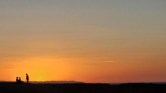 Drei Personen sitzen und stehen silhouettenhaft vor einem atemberaubenden Sonnenuntergang über den Sanddünen in der Mongolei.