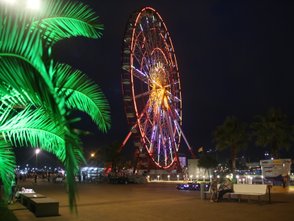 Ein großes, beleuchtetes Riesenrad steht im Hintergrund, während Menschen auf einer Bank im Vordergrund sitzen und sich unterhalten.