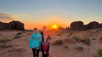 Drei Personen stehen im Monument Valley, während die Sonne hinter den Felsen untergeht und den Himmel in warmen Farben erleuchtet.