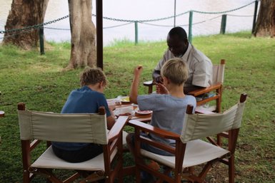 Zwei Kinder sitzen an einem Tisch im Freien und genießen eine Mahlzeit, während ein Erwachsener ihnen zuschaut.