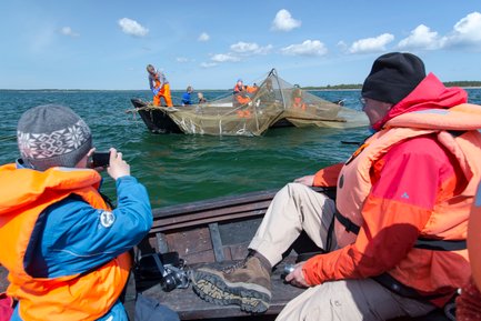 Ein Kind in einer orangefarbenen Schwimmweste fotografiert eine Gruppe von Menschen, die ein Netz auf einem Boot auswerfen.