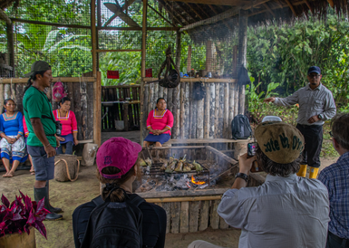 Eine Gruppe von Menschen sitzt um ein Feuer in einer traditionellen Hütte, während ein Mann ihnen etwas erklärt.