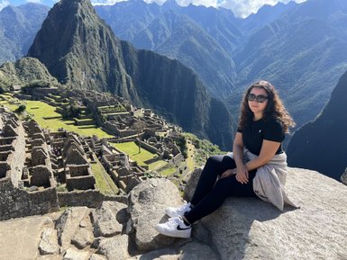 Eine Person mit lockigem Haar sitzt auf einem Felsen und blickt auf die beeindruckenden Ruinen von Machu Picchu.