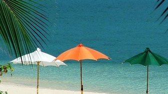 Drei Sonnenschirme in Weiß, Orange und Grün stehen am Strand, umgeben von feinem Sand und klarem, blauem Wasser.