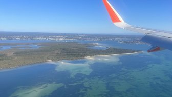 Das Bild zeigt den Blick aus einem Flugzeugfenster auf eine malerische Küstenlinie und glitzerndes Wasser.