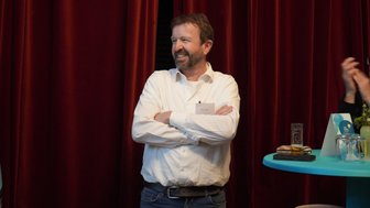 Ein Mann mit Bart steht lächelnd mit verschränkten Armen vor einem roten Vorhang, während er an einem Event teilnimmt.