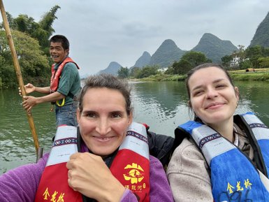 Zwei Frauen lächeln fröhlich in einem Bambusfloß, während ein Mann mit einem Paddel im Hintergrund steht.