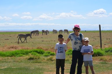Eine Frau steht mit zwei Kindern in einem grünen Feld, während Zebras im Hintergrund grasen und der Himmel blau ist.