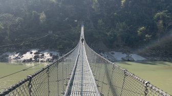 Eine schmale Hängebrücke erstreckt sich über den Modi Khola, umgeben von üppigem Grün und sanften Hügeln im Hintergrund.
