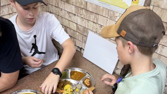 Zwei Jungen sitzen an einem Tisch und genießen indisches Streetfood von einem runden Metalltablett mit verschiedenen Gerichten.