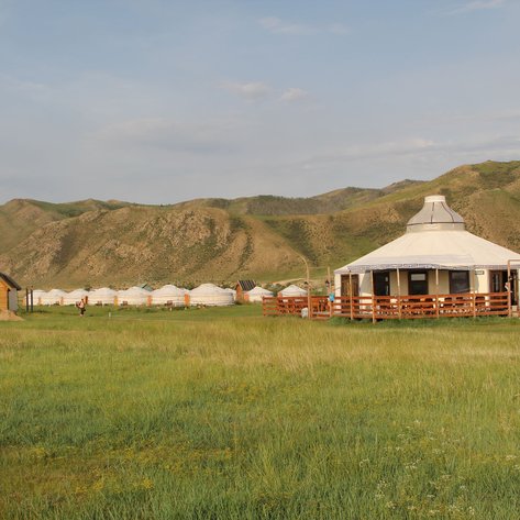 Eine malerische Landschaft mit Jurten und einem Holzhaus, umgeben von sanften Hügeln und grünem Gras.